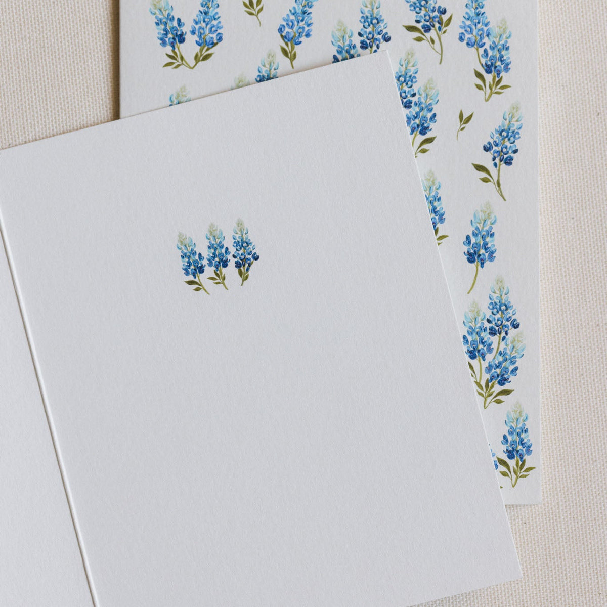 A white stationery card with a small blue and green floral print, layered over a matching patterned envelope on a textured beige fabric background.