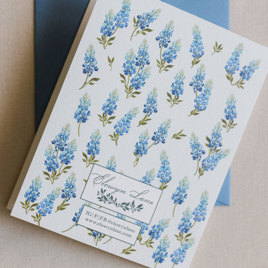 A square white invitation card with a repeating watercolor blue lupin floral pattern and a blue envelope behind it, photographed on a neutral beige surface.