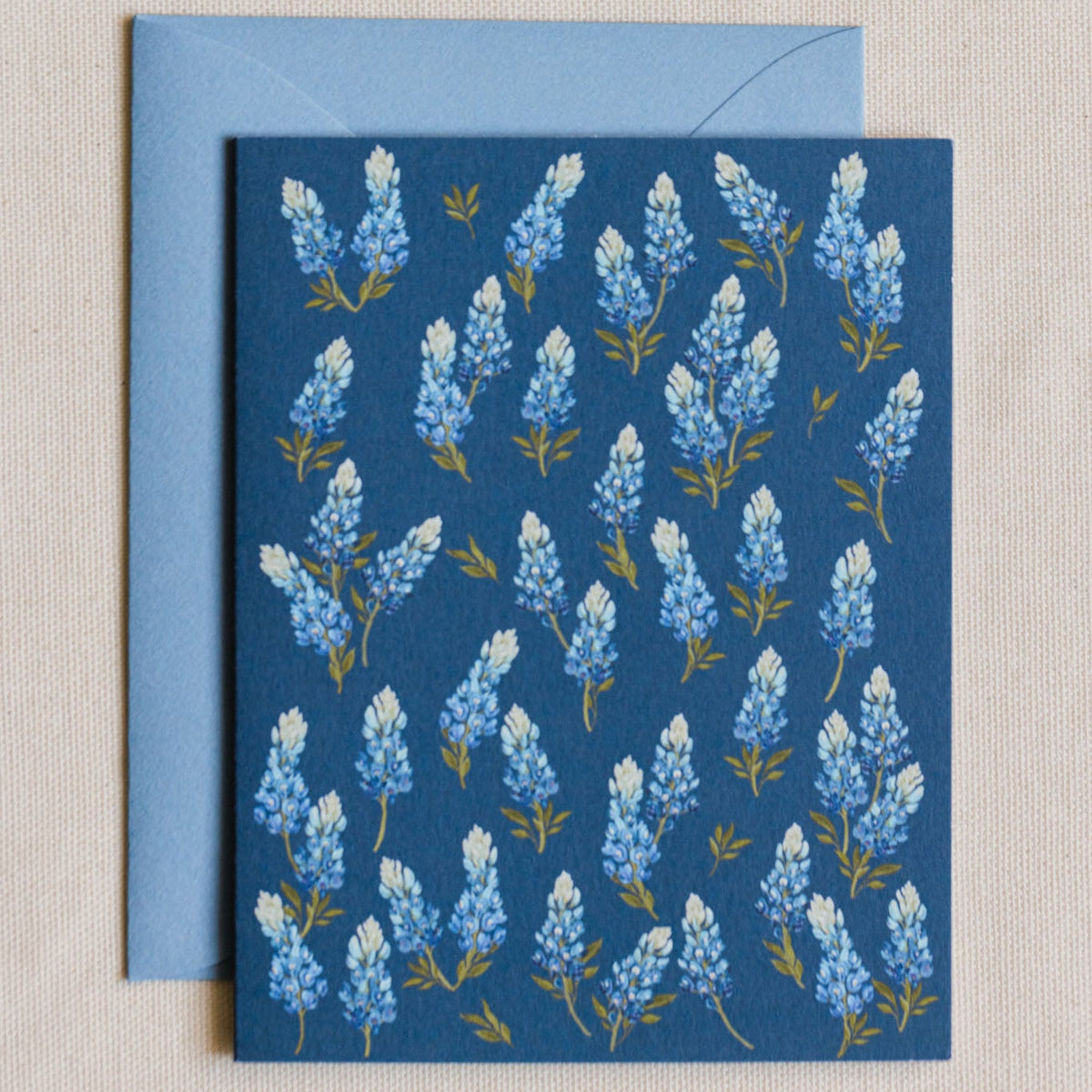 A square blue greeting card with a repeating pattern of white and light blue flowers with small yellow-green stems, shown with a light blue envelope on a beige textured fabric background.