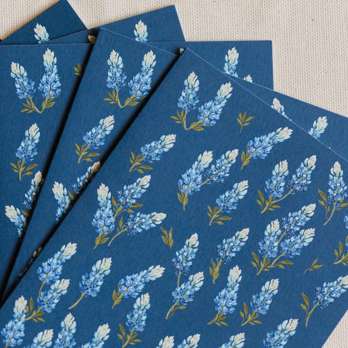 A stack of dark blue patterned cards or envelopes featuring a repeating print of light blue and white flowers with small yellow-green stems, arranged on a beige textured fabric background.