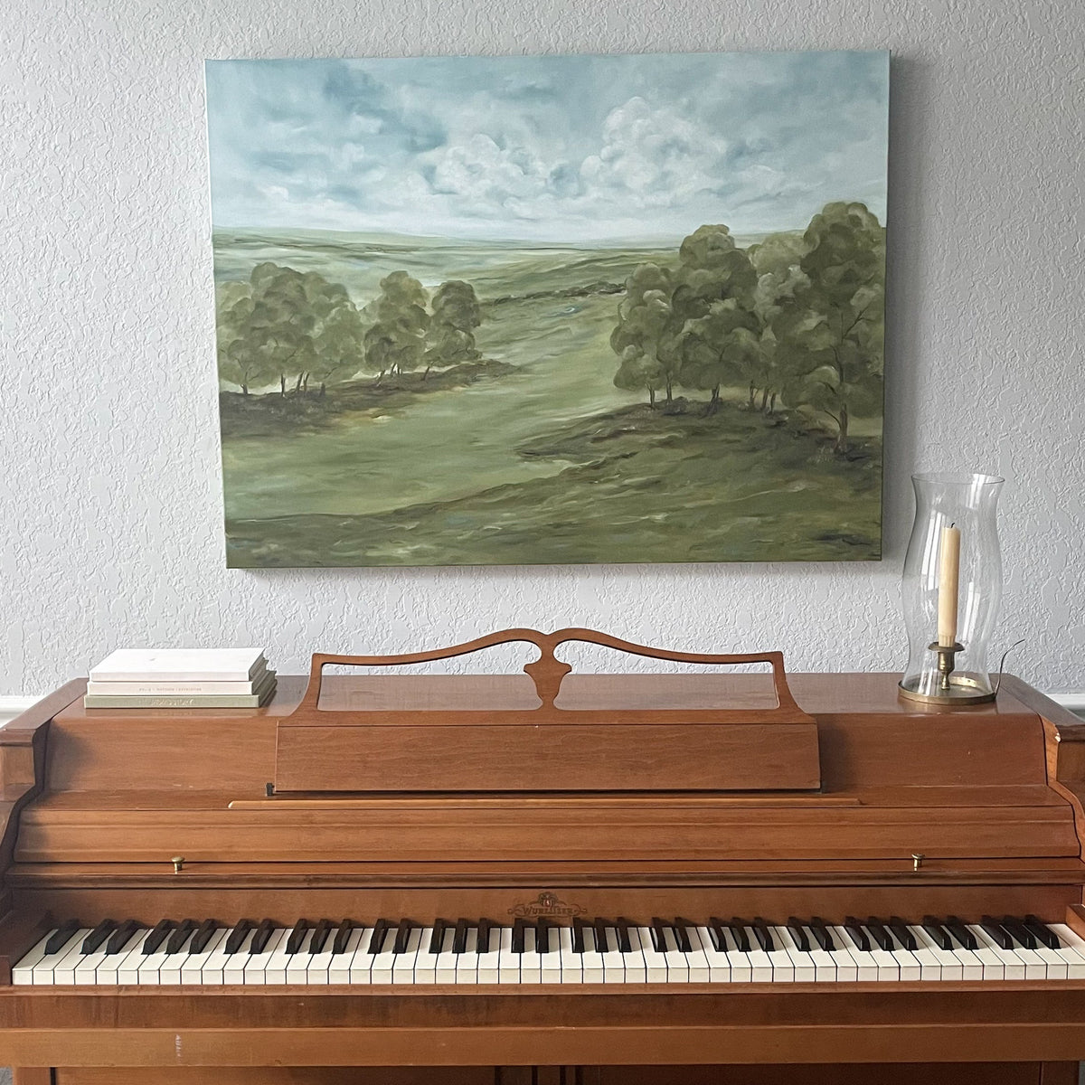 A landscape painting of rolling green hills and trees under a cloudy blue sky, displayed on a light gray wall above a wooden upright piano.