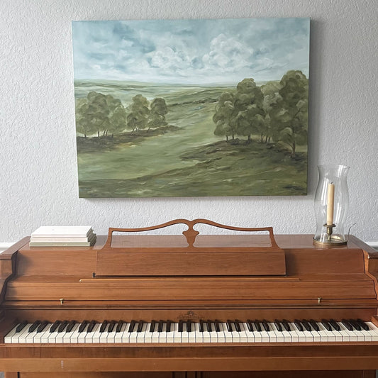 A landscape painting of rolling green hills and trees under a cloudy blue sky, displayed on a light gray wall above a wooden upright piano.