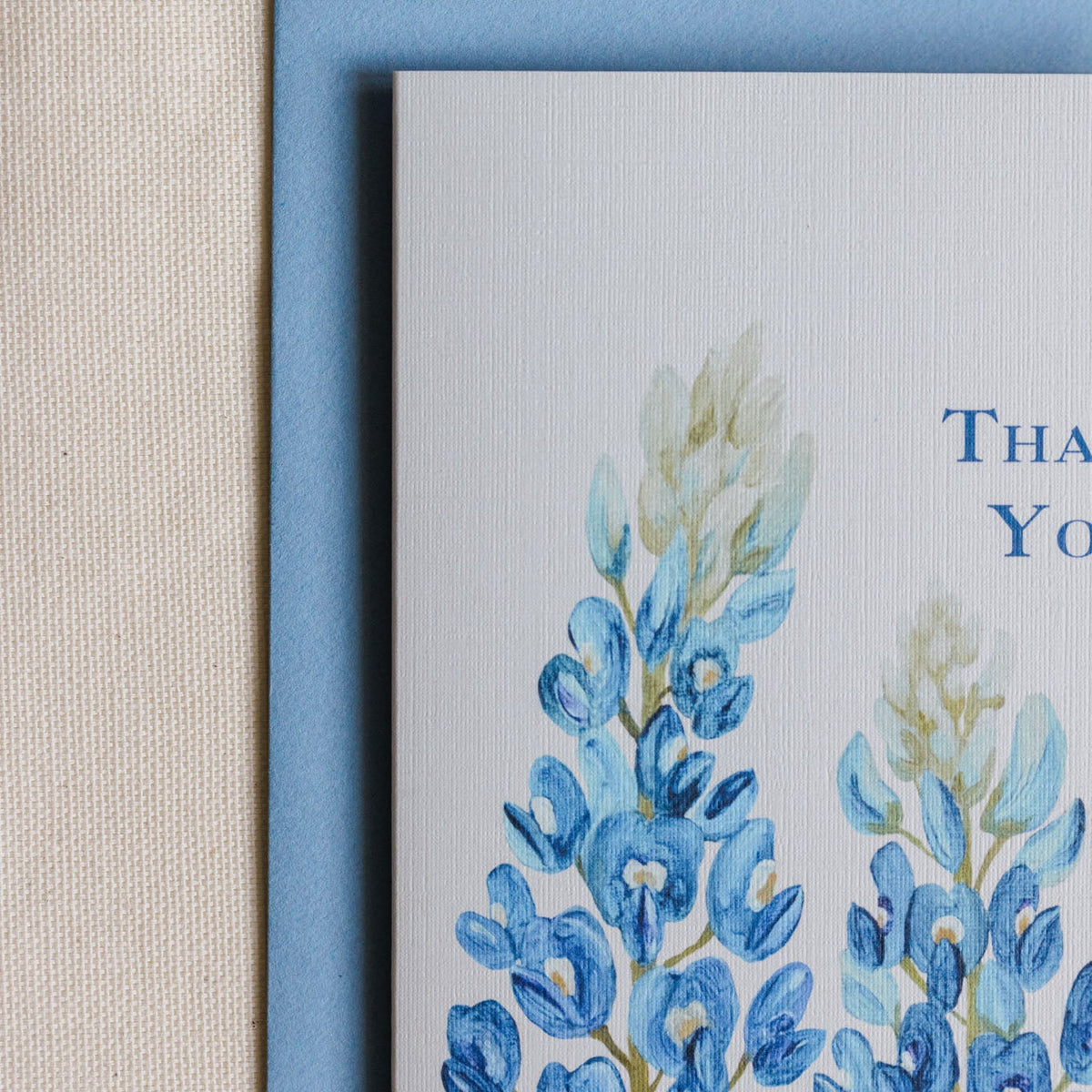 A close-up of a greeting card with a light blue envelope, featuring a watercolor illustration of blue lupines on a white card with partial blue text reading 'THA.