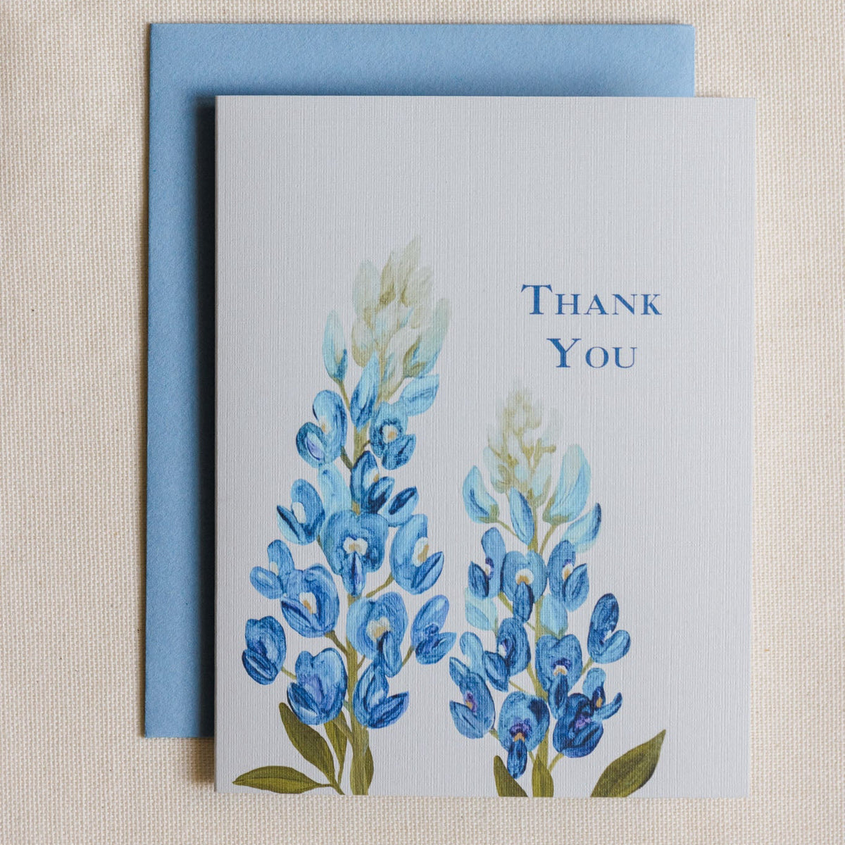 A square thank you card with a watercolor-style blue flower illustration and the words 'THANK YOU' on the right, placed in front of a light blue envelope on a beige fabric background.