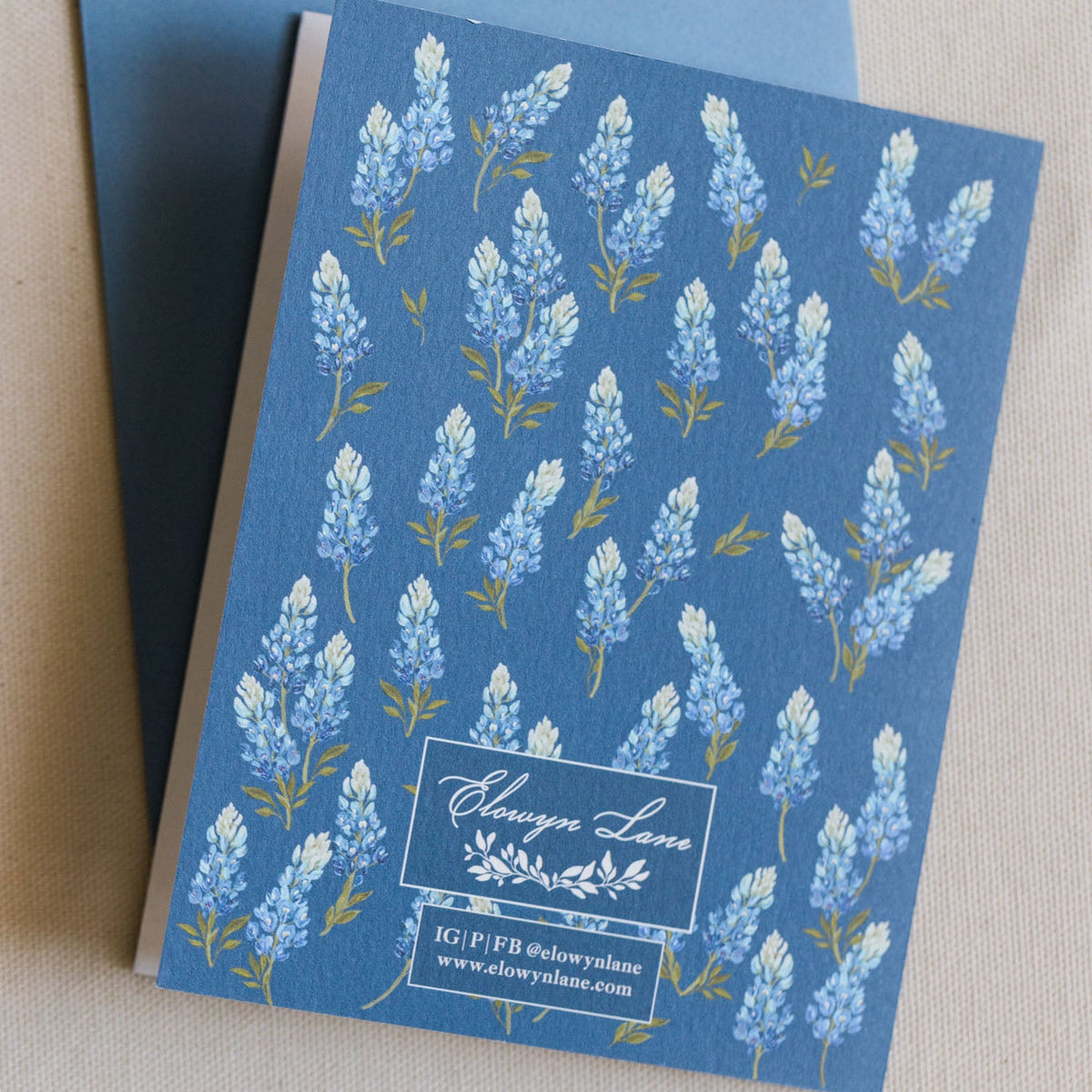 A blue greeting card with a white and light blue floral pattern and a matching blue envelope, displayed on a neutral beige textured background.