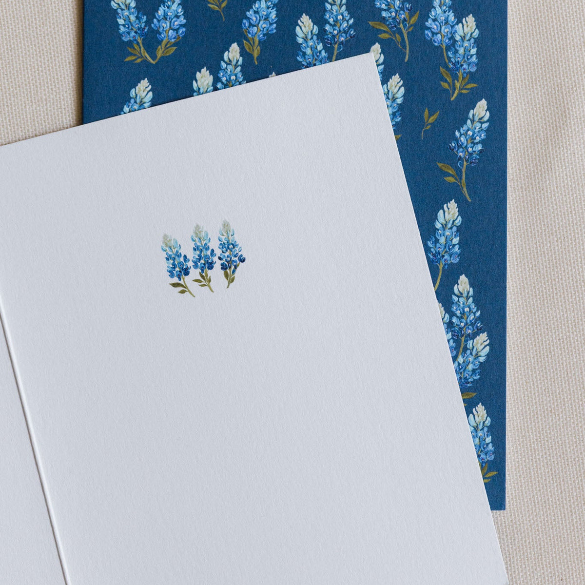 A white greeting card with a small blue and green floral illustration, layered over a navy blue envelope featuring a repeating blue and white floral pattern, set against a neutral beige textured background.