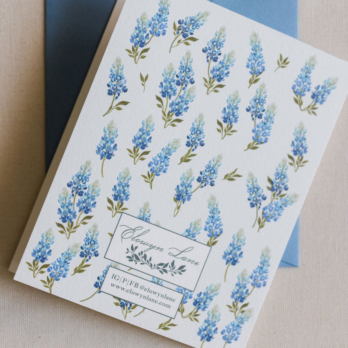 A square white invitation card with a repeating watercolor blue lupin floral pattern and a blue envelope behind it, photographed on a neutral beige surface.