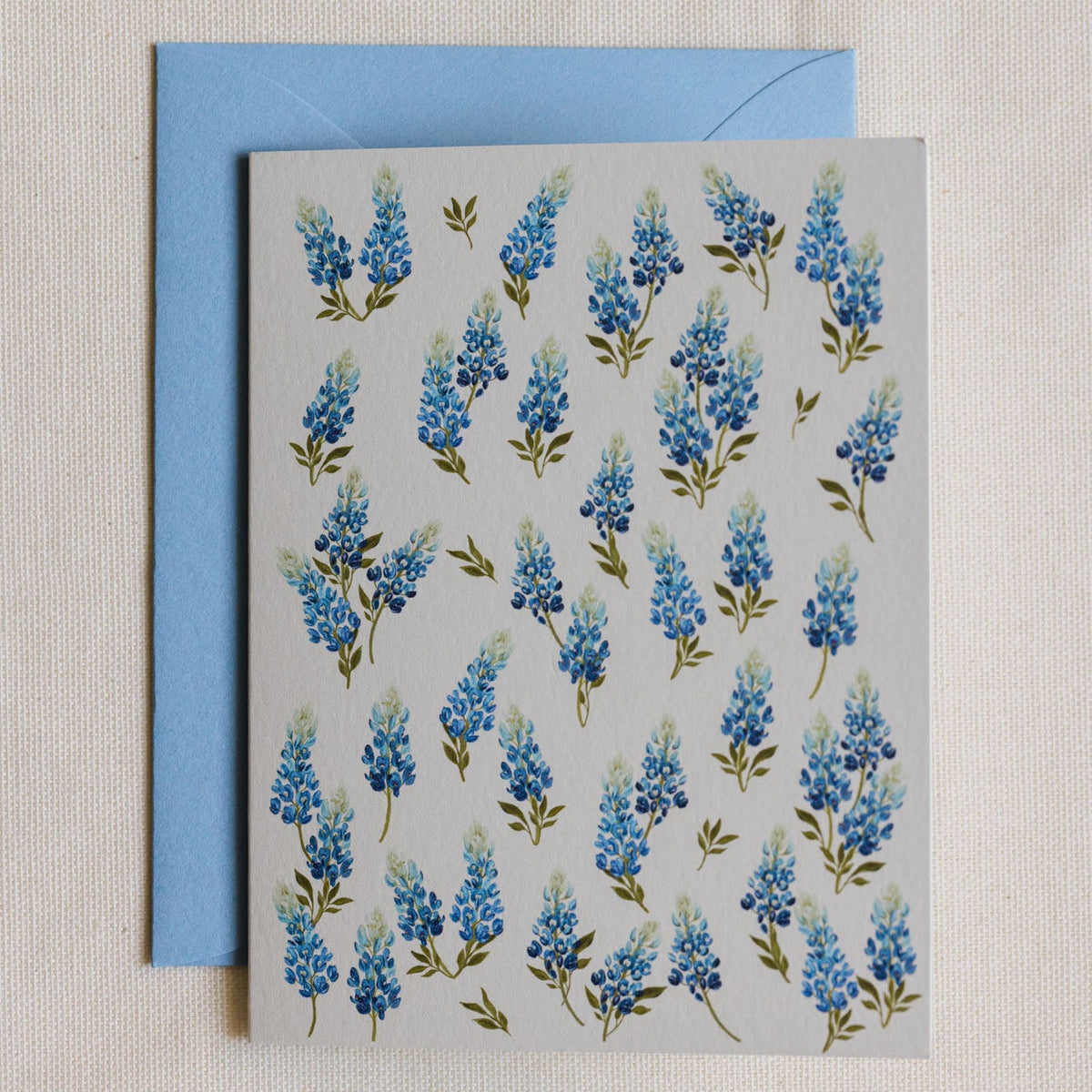 A square greeting card with a repeating blue flower pattern on a light gray background, shown with a light blue envelope behind it on a textured cream fabric surface.