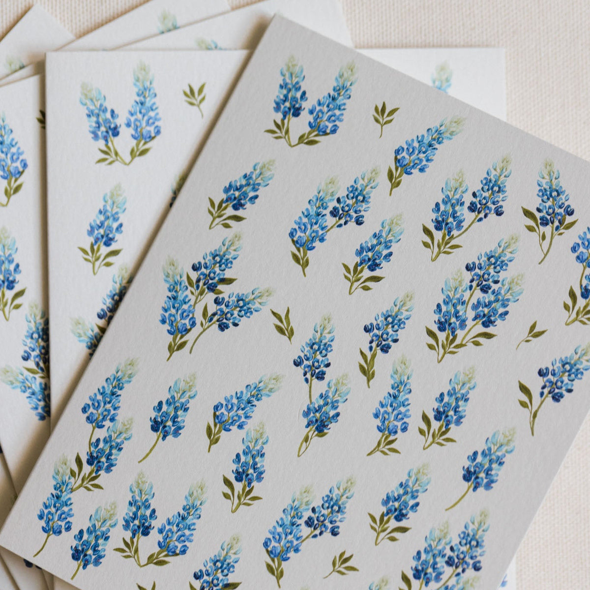 A stack of white greeting cards featuring a repeating watercolor-style blue flower and green leaf pattern, arranged on a light beige textured fabric background.