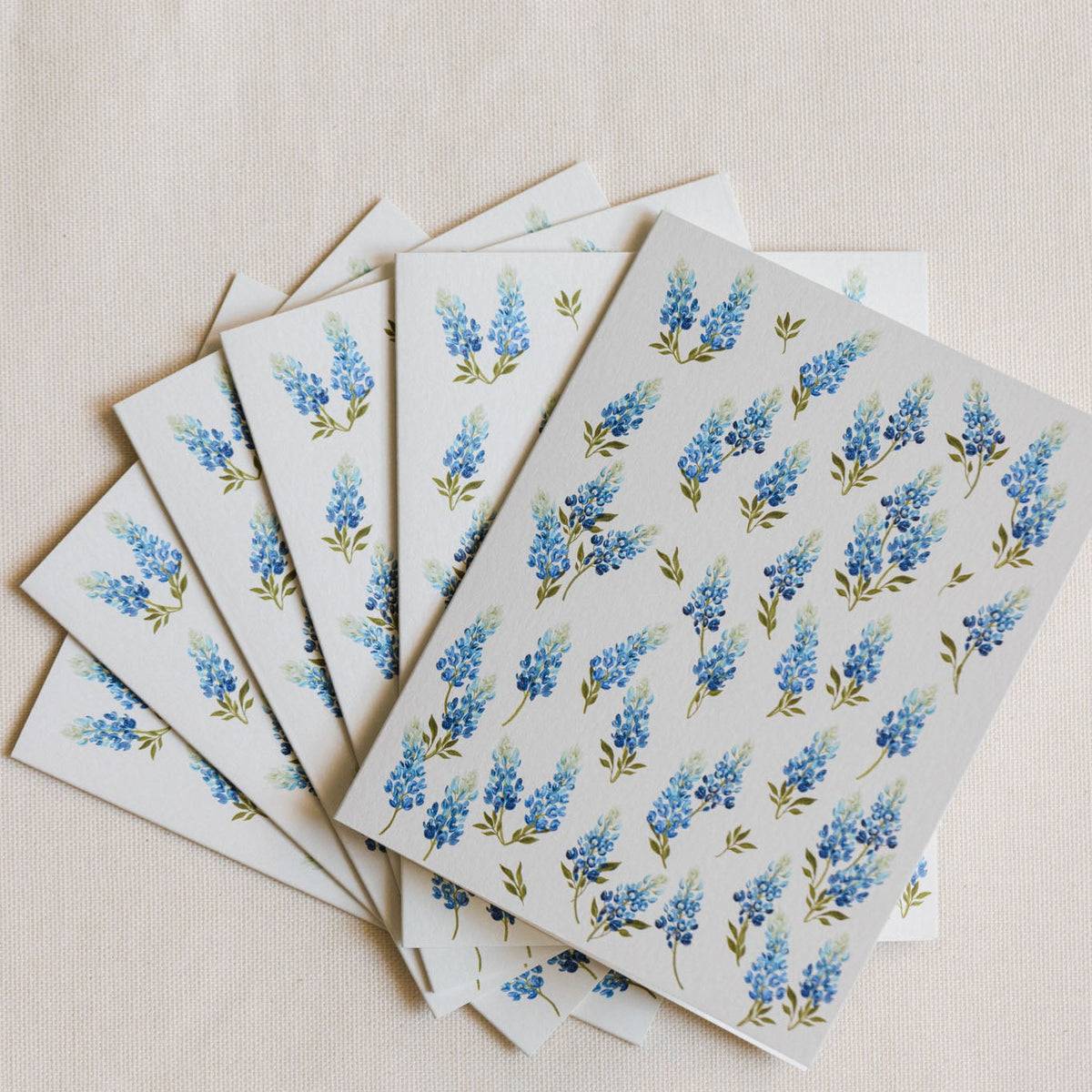 A stack of white rectangular cards or envelopes printed with a repeating blue flower and green leaf pattern, arranged in a fan shape on a light beige textured background.