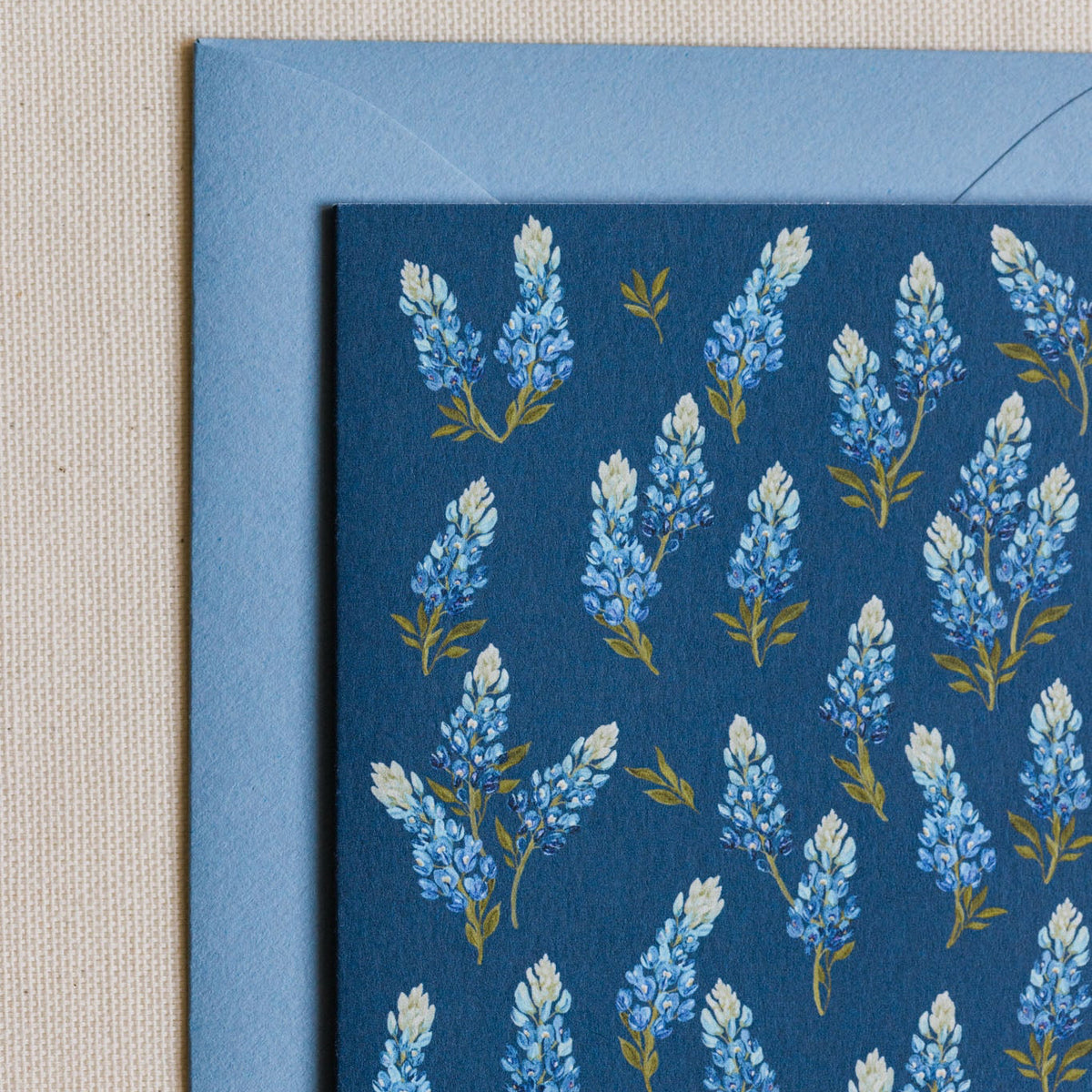A close-up of a square card or paper product with a blue floral pattern, layered over a light blue envelope on a beige textured fabric background.