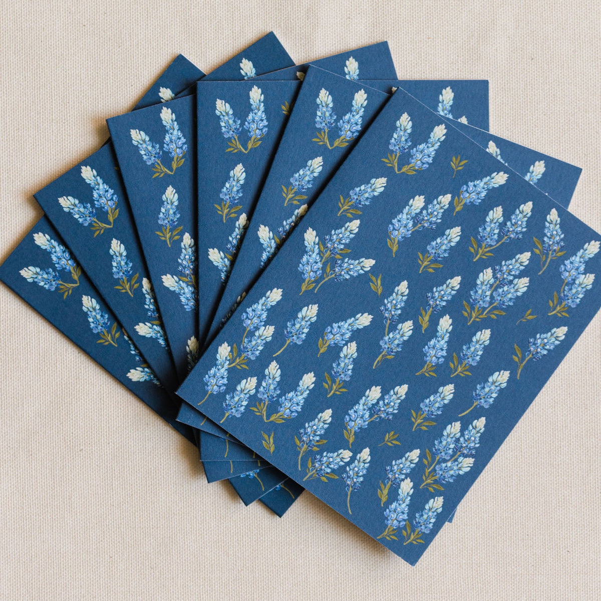 A fan arrangement of blue paper napkins or cards featuring a repeating white and light blue floral pattern with small gold accents, laid on a light beige fabric background.
